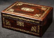 JB506:  Rosewood box  with rounded edges, inlaid with brass to the front and top depicting stylized flora.  The design is controlled in deference to the Georgian neo-classical tradition but it also embraces the Regency influence of the Royal cabinet maker George Bullock, who introduced naturalistic elements to the earlier austere brass designs.  The inside of the box has been relined with hand marbled paper and velvet  and retains  its original lift out tray.  Circa 1825.  Enlarge Picture