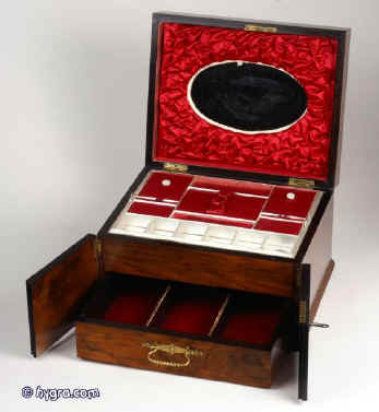 JB303: A figured walnut table cabinet with double doors to the front opening to reveal a drawer fitted for jewelry The box/cabinet also has  a compartmentalized lift out tray with supplementary lids covered in red satin.  There is an oval mirror on the inside of the lid framed with ruched red satin. Circa 1890. Enlarge Picture
