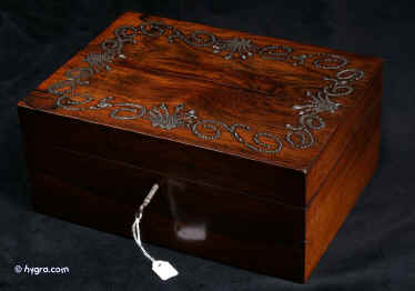 JB405: Early 19th century box veneered in dark figured rosewood and decorated with cut steel pins. The steel was cut in the manner of precious stones and on this box it was arranged in a neoclassical pattern of symmetrical scrolls centred by a palmette motif. Cut steel work was popular for a short period of time during the late 18th century and the early 19th century. The work which is laboriously slow needs precision and skill of very high calibre. The box has been fitted with a lift-out tray. The box has a working lock and key. Crack on top veneer. Circa 1815.Enlarge Picture