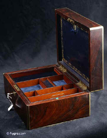 JB407: William IV/early Victorian box veneered in strongly figured rosewood and edged in rounded brass, which defines the lines and strengthens the structure. Inside the original tray has been fitted with additional velvet pads. The flap on the inside top lid retains the original leather complete with fine gold embossed decoration in the neoclassical tradition. It opens down to an 'envelope' lined in original pink paper. Veneer cracking on top not affecting the structure. The box has a working lock and key. Circa 1835.  Enlarge Picture