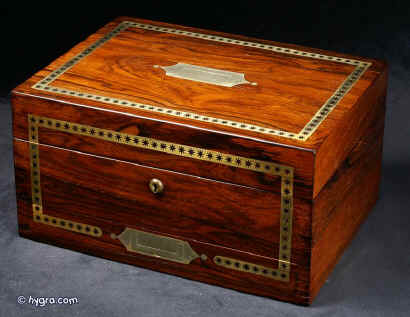 JB408: Regency box veneered in figured rosewood and decorated with strips of brass with cut out star motifs. Symmetrical restrained decoration of this type is typical of the early part of the 19th century. The box has a lower pull-out drawer with a central brass handle. The top part interior has been separated and lined. The leather cover on the envelope compartment on the inside top lid is original and it is embossed in a neoclassical design incorporating anthemia and palmettes. When it is folded down it reveals a mirror. The mirror is a new addition, although it sits in the alcove originally intended for a mirror. Some cracking to top veneer. The box has a working lock and key. Circa 1815. Enlarge Picture