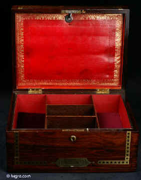 JB408: Regency box veneered in figured rosewood and decorated with strips of brass with cut out star motifs. Symmetrical restrained decoration of this type is typical of the early part of the 19th century. The box has a lower pull-out drawer with a central brass handle. The top part interior has been separated and lined. The leather cover on the envelope compartment on the inside top lid is original and it is embossed in a neoclassical design incorporating anthemia and palmettes. When it is folded down it reveals a mirror. The mirror is a new addition, although it sits in the alcove originally intended for a mirror. Some cracking to top veneer. The box has a working lock and key. Circa 1815.  Enlarge Picture