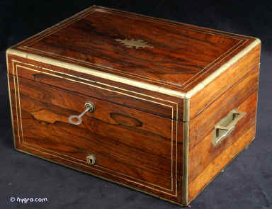 JB410: Early 19th century box veneered with exceptionally beautifully figured rosewood. The box is edged in rounded brass and further enhanced with two lines of brass stringing. It has brass handles to the sides. There is a lower drawer with a separate lock to the front. This is divided in three compartments. The top part of the box has a lift-out tray and a flap-down envelope compartment. Both retain the original lining of green leather and velvet. The maker's label is visible stuck on the inside of the flap. The lower inside of the box is also covered in the original leather. Small veneer cracks to the top. The box has two working locks and one key which opens both locks. Circa 1820. Enlarge Picture