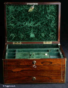 JB410: Early 19th century box veneered with exceptionally beautifully figured rosewood. The box is edged in rounded brass and further enhanced with two lines of brass stringing. It has brass handles to the sides. There is a lower drawer with a separate lock to the front. This is divided in three compartments. The top part of the box has a lift-out tray and a flap-down envelope compartment. Both retain the original lining of green leather and velvet. The maker's label is visible stuck on the inside of the flap. The lower inside of the box is also covered in the original leather. Small veneer cracks to the top. The box has two working locks and one key which opens both locks. Circa 1820.  Enlarge Picture