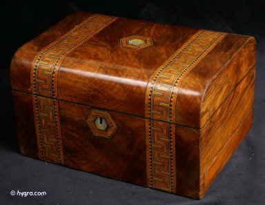 JB412: Victorian box veneered in figured walnut and decorated with straps of geometric inlay in a Greek pattern. The inlay is executed in ebony and fruitwoods with the wide strips in cross-cut kingwood. The box has a sprung side drawer with two partition strips covered in the original leather. The top part has the original lift-out tray to which velvet covers have been added to the bottom parts of the partitions. The inside of the lid has a spring out mirror which can be clicked in place either on the mirror or the velvet covered side. The velvet is original as is the leather framing on both the mirror and the ruched velvet. This is further enhanced with a delicate line and corner motif decoration in gold. The escutcheon and central plaque are centred in mother of pearl. Working lock and key. Minute cracks and tiny corner repairs. Circa 1875. Enlarge Picture