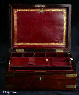 JB414: Early Victorian box veneered in rosewood and bound with brass straps and unusually shaped brass corners which soften the formality of the brass with a gently rounded curve. The interior and the lift-out tray are covered in the original wine-coloured leather. Under the tray the space is further compartmentalised with original leather covered separations. The back lid envelope pouch is again covered in the original leather and decorated with a gold embossed pattern of palmettes. Working lock and key. Circa 1835-40. Enlarge Picture