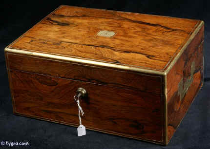 JB418: Early 19th century box veneered in exceptionally strongly figured rosewood. The box is edged in rounded brass and further enhanced with a line of brass stringing and elegant side handles. The inside facings of the box are also enhanced with fine brass stringing. The hinge is most unusual, of a rounded form reminiscent of the Scottish hinge structure. The interior retains its original leather covered tray which has had velvet covers added to the bottom of the sections. A removable velvet cover has also been made for the back lid. Working Bramah lock and key. Circa 1820. Enlarge Picture