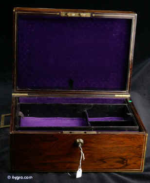  JB418: Early 19th century box veneered in exceptionally strongly figured rosewood. The box is edged in rounded brass and further enhanced with a line of brass stringing and elegant side handles. The inside facings of the box are also enhanced with fine brass stringing. The hinge is most unusual, of a rounded form reminiscent of the Scottish hinge structure. The interior retains its original leather covered tray which has had velvet covers added to the bottom of the sections. A removable velvet cover has also been made for the back lid. Working Bramah lock and key. Circa 1820. Enlarge Picture
