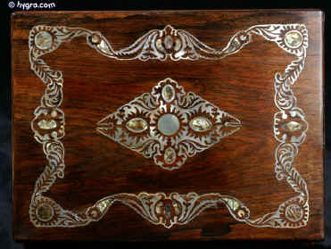 JB419: Mid Victorian box veneered in dark rosewood and decorated with mother of pearl. The pattern of the decoration is controlled and symmetrical but it also swirls in the more naturalistic style of the later 19th century. The motifs are of stylised flora which is accented with centres of darker abalone shell. The box has a spring-out side drawer covered in original silk. The interior has been fitted with another tray. The back lid retains its original drop down envelope compartment covered in original ruched velvet.  Small repairs and minute losses to mother of pearl visible under close scrutiny. The box has a working lock and key. Circa 1870. Enlarge Picture