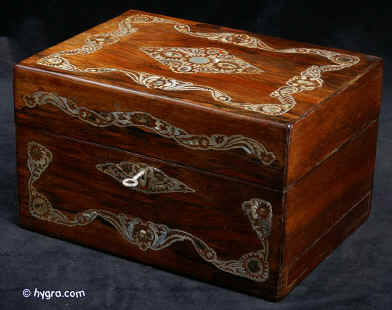 JB419: Mid Victorian box veneered in dark rosewood and decorated with mother of pearl. The pattern of the decoration is controlled and symmetrical but it also swirls in the more naturalistic style of the later 19th century. The motifs are of stylised flora which is accented with centres of darker abalone shell. The box has a spring-out side drawer covered in original silk. The interior has been fitted with another tray. The back lid retains its original drop down envelope compartment covered in original ruched velvet.  Small repairs and minute losses to mother of pearl visible under close scrutiny. The box has a working lock and key. Circa 1870. Enlarge Picture