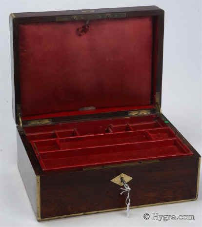 JB446: Figured rosewood and brass surround box circa 1830. A box veneered in figured rosewood box strengthened and accented with brass. The top central plaque bears initials in a stylized Gothic  script and a coronet,  probably of French nobility. There is also a brass inset of a lift-up aid on the front part of the lid. The box has extra support hinges on the sides which enable it to stay open at the same angle. The interior tray is original as is the silk faced envelope. Additional velvet covers protect and render the interior more useable. Working key. Enlarge Picture