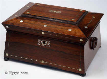 Rosewood veneered box in neoclassical form circa 1835. A box inspired by the shapes of classical temples fashionable in the second phase of neoclassicism. The structure with its angled and tiered sections gives the impression of elegance and strength. The decoration in mother of pearl and white metal is restrained. It further emphasizes the strength of the lines and enlivens the colour of the wood. The escutcheon and central motif in controlled stylized flora give a note of softness. The original lift-out tray has been re-covered in cotton velvet. The inside of the lid is covered in the original ruched silk surrounded by gold-embossed leather. The handles are original albeit with some old breaks and repairs. The feet are not original. Working lock and key. Enlarge Picture