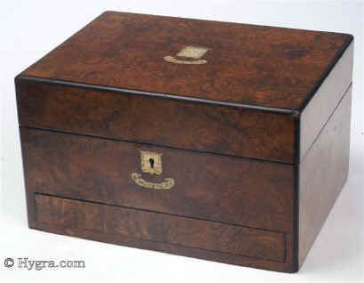 JB461 Burr walnut veneered box circa 1870. A box veneered in strikingly figured burr walnut. The central plaque and motif are of engraved brass. There is no further decoration, the attraction being the very pretty figure of the wood. The box has a lower drawer which opens with a spring mechanism. On the top part, the box has a later lift out tray. The box has been relined. The mirror, which clicks in place on the inside top part, is original as is its embossed leather framing. The box has small veneer repairs and some damage above the lock. Working lock and key. Enlarge Picture