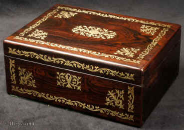 JB506:  Rosewood box  with rounded edges, inlaid with brass to the front and top depicting stylized flora.  The design is controlled in deference to the Georgian neo-classical tradition but it also embraces the Regency influence of the Royal cabinet maker George Bullock, who introduced naturalistic elements to the earlier austere brass designs.  The inside of the box has been relined with hand marbled paper and velvet  and retains  its original lift out tray.  Circa 1825. Enlarge Picture