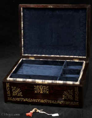 JB506:  Rosewood box  with rounded edges, inlaid with brass to the front and top depicting stylized flora.  The design is controlled in deference to the Georgian neo-classical tradition but it also embraces the Regency influence of the Royal cabinet maker George Bullock, who introduced naturalistic elements to the earlier austere brass designs.  The inside of the box has been relined with hand marbled paper and velvet  and retains  its original lift out tray.  Circa 1825.Enlarge Picture