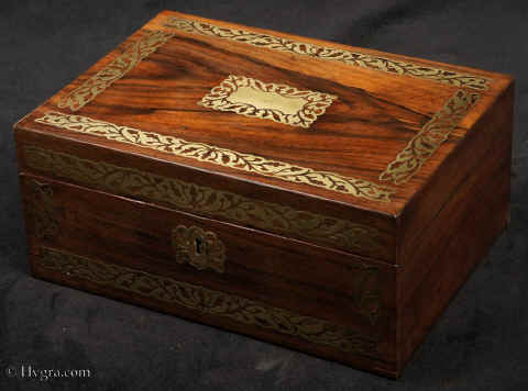JB507: Rosewood box inlaid with brass to the front and top depicting stylized flora.  The design is controlled in deference to the Georgian neo-classical tradition but it also embraces the Regency influence of the Royal cabinet maker George Bullock, who introduced naturalistic elements to the earlier austere brass designs. The inside of the box has been relined with hand marbled paper and velvet  and retains  its original lift out tray.  Circa 1825. Enlarge Picture