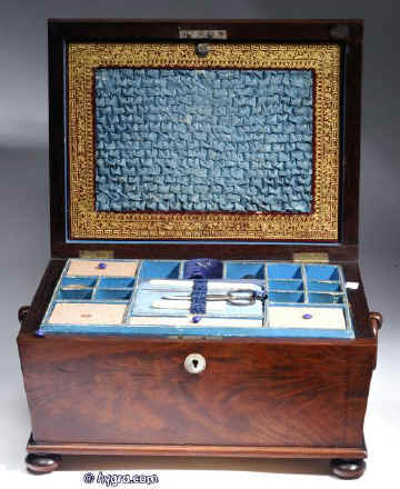 SB126: Antique flame mahogany sewing box with tapered sides and pyramided top having turned mahogany drop handles and standing on turned bun feet. Inside the lid is lined with ruched blue silk framed with gold embossed leather. There is a document wallet behind. The box has a fitted liftout tray covered in the original blue paper and has silk covered supplementary covers circa 1815.  Enlarge Picture