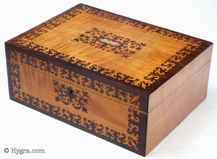  Sb451: Satinwood and rosewood veneered box with stylised floral design circa 1835. A beautifully elegant box. The composition contrasts the rich brown of the rosewood with the luminous sheen of the satinwood in stylized floral motifs, inspired both by earlier seaweed patterns and later neoclassical designs. The inlay is executed with impeccable precision and control and becomes an integral part of the whole structure of the box. The top central plaque is in mother of pearl and gives an extra punctuation to the composition. Small repairs to two of the corners on the top, seen when carefully examined. The tray is original and so is the blue paper and metal bead edging on the tray. The velvet covers are re-covered. Working lock and key. Enlarge Picture