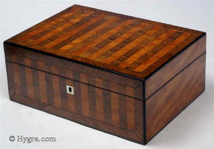 SB453: Satinwood and rosewood box veneered in stripes circa 1840. This is a fully fitted sewing box which oozes quiet elegance. The stripes emphasise the contrasting colours and varied figures of the two special timbers which cover the top and front. The box is edged in rounded rosewood giving the form a neat and defining line. The tray is edged in ebony, an unusual detail which contributes both to beauty and resistance to wear. The blue silk is original and so is the envelope under the lid. There is a set of seven hand made mother of pearl spools with metal shafts. This form of spool became fashionable in the 1830s when mercerised thread became increasingly more available. The tray also contains other mother of pearl and steel sewing tools. Working lock and key. Enlarge Picture