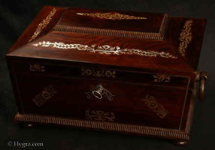SB487: Antique  figured rosewood sewing box the top panel and base  framed by gadrooning, inlaid with mother of pearl to the top and front having turned rosewood drop-ring handles and standing on  turned bun  feet. Inside the lid is lined with  cream silk and paper. Inside the lid there is a mirror framed by gold embossed leather with a document wallet behind The box has a liftout tray with supplementary lids covered in silk. Circa 1830. Enlarge Picture