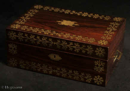 SB524: Rosewood and brass inlaid sewing box in high Regency style circa 1815.  A box veneered in beautifully figured rosewood and inlaid with brass. The design of the inlay is of highly stylized flora, suggesting neoclassical designs hinting at trefoil motifs. The juxtaposition of dark wood with bold brass inlay was popular in the early part of the 19th century. The wood and the bright brass were mutually enhancing. The box retains its original  fully fitted tray covered in yellow paper with gold embossed supplementary lids. There is a set of  eight turned and carved vegetable ivory  spools. Enlarge Picture