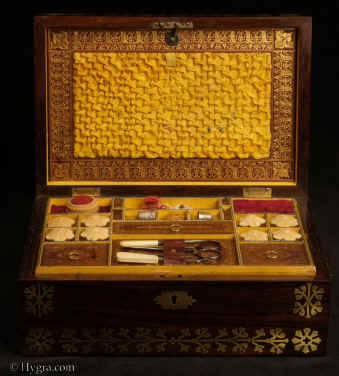 SB524: Rosewood and brass inlaid sewing box in high Regency style circa 1815. A box veneered in beautifully figured rosewood and inlaid with brass. The design of the inlay is of highly stylized flora, suggesting neoclassical designs hinting at trefoil motifs. The juxtaposition of dark wood with bold brass inlay was popular in the early part of the 19th century. The wood and the bright brass were mutually enhancing. The box retains its original  fully fitted tray covered in yellow paper with gold embossed supplementary lids. There is a set of  eight turned and carved vegetable ivory  spools. Enlarge Picture