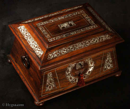TC566:Monumental three canister tea chest in figured rosewood on a mahogany carcass with mother of pearl inlay standing on turned rosewood feet having turned rosewood drop ring handles. The shape of this chest combines elements and influences characteristic of the  nineteenth century. It is structured in an   ancient Egyptian architectural form combining tapered  and pyramid lines which make this caddy a strong statement of the robust and elegant style of the Regency. The composition of the top is strong. The inlaid mother of pearl inlay  panels are set off by a further stringing of white-metal. (pewter). circa 1830.  Enlarge Picture