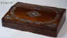 WB441: Writing slope in rosewood and satinwood (inside) with mother of pearl inlay circa 1835. This is a writing slope of exceptionally high quality. The outside is veneered in saw cut rosewood inlaid with mother of pearl. The inlay is executed with precision in two different ways: The central and corner parts have the floral patterns outlined in rosewood with the mother of pearl surrounding them; the main encircling part is made out of mother of pearl inlaid into the rosewood. On examining the rosewood patterns closely, one can see fine lines separating the leaves, a sure sign that the box retains its original finish. The design is both elegant and robust, straddling the time between the neoclassical and the naturalistic period. The patterns are symmetrical and rendered with control of line and space. This shows the neoclassical influence. There are also neoclassical motifs such as the anthemion, acanthus leaves and palmettes. However the flowers, although symmetrically arranged and stylised, have more than a hint to naturalistic aspirations and the neoclassical motifs are integrated within the whole composition. The garland, although controlled and defined within a perfect oval, loops and curls in fluid movement like a living trellis. Inside the box has a pen tray and lift up lids. The underside of the lids has at some stage been stained. There is one original inkwell with a silver plated screw on top. The writing surface is covered in subtly embossed velvet, which we think is a later replacement, but very close to what a box of this period cud have had.  The interior of the box continues the high standard of quality and it is veneered in satinwood. Small crack/repair on the top part of the veneer, perceptible when the box is closely examined.  Working lock and key.Enlarge Picture